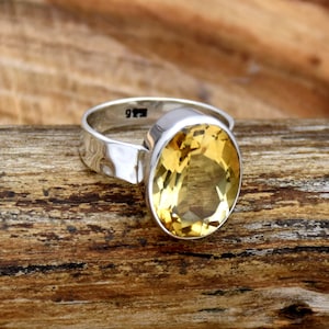 May include: A silver ring featuring a large oval-shaped yellow citrine gemstone. The ring is displayed on a wooden surface.