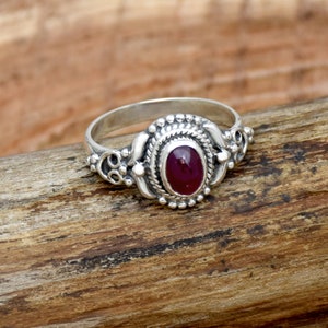 Puede incluir: Un anillo de plata con una piedra preciosa ovalada de color rojo oscuro engastada en un diseño detallado y ornamentado.
