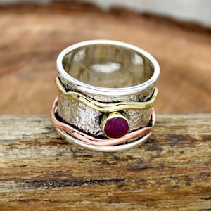 May include: A silver ring with a red gemstone set in the center. The ring has three bands of different metals: silver, gold, and copper. The bands are textured and have a wavy design.