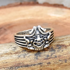 May include: A silver ring featuring a detailed, stylized mask design. The ring has a band with horizontal lines and a raised mask with intricate facial features. The ring is set against a natural wood background.