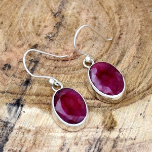 May include: A pair of silver dangle earrings with oval-shaped ruby gemstones. The earrings have a simple design and are perfect for everyday wear.