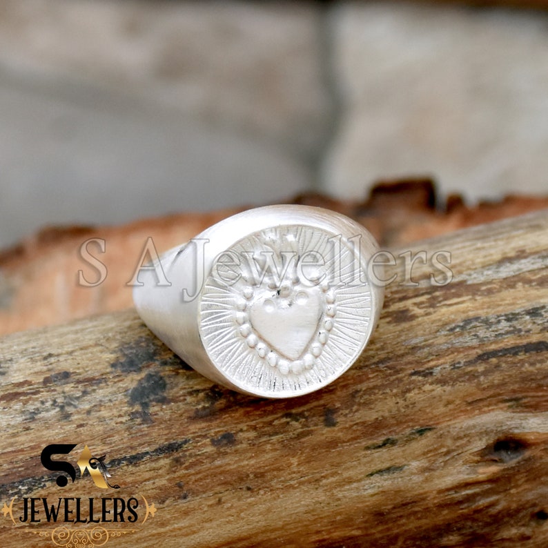 May include: A silver ring with a heart design. The heart is surrounded by a circle of small beads and rays of light. The ring is on a wooden surface.