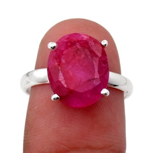 Pode incluir: Um anel de prata com uma grande gema de rubi oval. O rubi é de uma cor vermelho-rosa vibrante, preso por quatro garras de prata. A banda do anel é de prata polida.