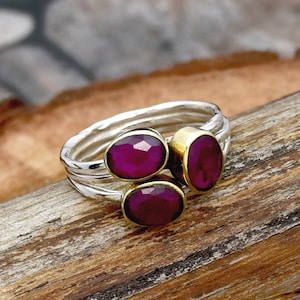 May include: A silver ring featuring three oval-shaped, faceted deep pink gemstones. Two of the stones are set in gold-toned bezels, while the third is set in a silver-toned bezel. The band has a textured, layered appearance with a metallic finish.
