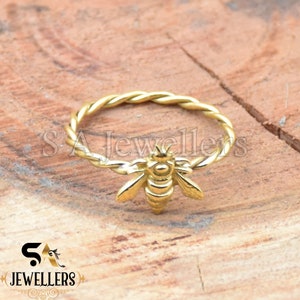 Può includere: Un anello dorato con una fascia intrecciata e un design a forma di ape. L'ape è rivolta in avanti con le ali spiegate.