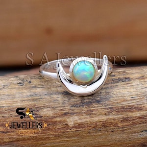May include: A silver ring featuring a crescent moon design cradling a round, iridescent opal. The opal displays a range of colors, including blue, green, and white. The ring is set against a wooden surface.