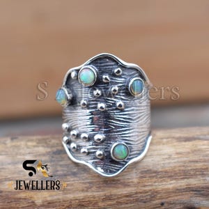 Op de afbeelding: Een brede, getextureerde zilveren ring met vier opaal edelstenen. De ring heeft een uniek, organisch ontwerp met kleine, verhoogde stippen en een gebeeldhouwd oppervlak. De opaalstenen zijn rond en hebben een subtiele, iriserende gloed. De ring wordt gepresenteerd op een houten oppervlak.