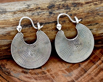 May include: A pair of silver hoop earrings with a circular design featuring concentric circles. The earrings are displayed on a wooden surface.
