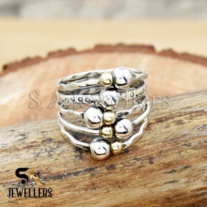 May include: A set of five silver rings with textured bands and gold beads. The rings are stacked on top of each other.