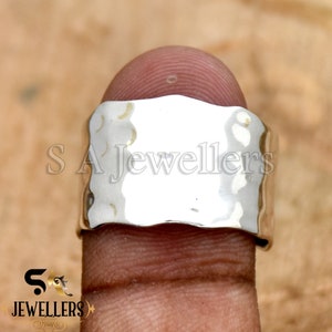 May include: A silver hammered ring with a wide band. The ring is on a finger.