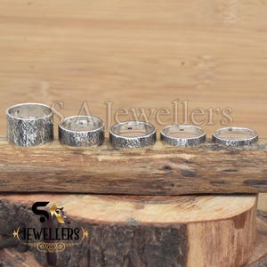 K&ouml;nnte beinhalten: F&uuml;nf silberne Ringe mit einer strukturierten Oberfl&auml;che, die nach Gr&ouml;&szlig;e auf einer Holzoberfl&auml;che angeordnet sind. Die Ringe sind alle im gleichen Stil, mit einem breiten Band und einer leicht abgerundeten Kante.