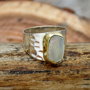 Op de afbeelding: Een zilveren ring met een gehamerde textuur en een gouden rand. De ring heeft een grote, rechthoekige, iriserende steen. De ring ligt op een houten oppervlak.