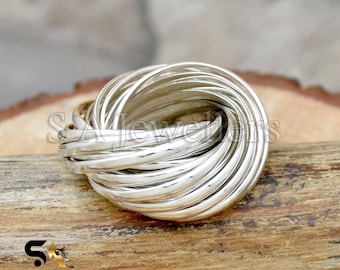 Könnte beinhalten: Ein silberner Ring aus mehreren ineinander verschlungenen Bändern, die ein knotenartiges Design bilden. Der Ring wird auf einer Holzoberfläche gezeigt.