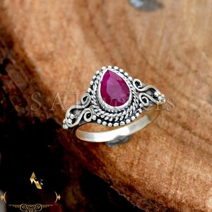 Può includere: Un anello d'argento con un designo ornato che presenta una pietra preziosa di rubino a forma di lacrima.