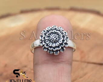 Könnte beinhalten: Ein Sterling Silber Ring mit einem detaillierten Blumenmuster. Der Ring hat eine Blume mit geschichteten Blütenblättern, die eine strukturierte, antike Oberfläche aufweist. Der Ring wird auf einem Finger vor einem verschwommenen Hintergrund präsentiert.