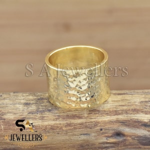 May include: A wide, hammered gold ring. The ring has a textured surface and a cylindrical shape. The ring is displayed on a wooden surface. The text "SA Jewellers" is visible in the background.