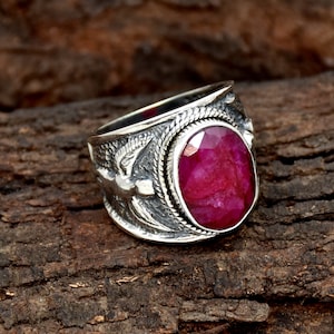 May include: A silver ring featuring a large, oval-shaped ruby gemstone. The ring has intricate detailing with a bird design on the band.