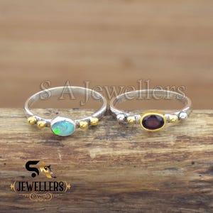 May include: Two silver rings with gemstone settings. One ring features an oval, light blue opal, and the other has a dark red oval gemstone. Both rings have gold and silver accents. The rings are displayed on a wooden surface.