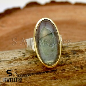 Puede incluir: Un anillo de plata con un engaste de oro que presenta una gran piedra de labradorita de forma ovalada. El anillo se muestra sobre una superficie de madera.