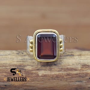 May include: A two-tone ring with a rectangular, deep red gemstone set in a gold-colored bezel. The band is silver-colored, with gold-colored accents on the sides. The ring is displayed on a wooden surface. The text "SA Jewellers" is visible.