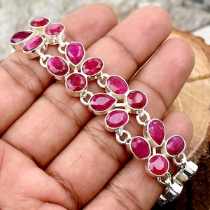 Könnte beinhalten: Ein silbernes Armband mit mehreren ovalen und tropfenförmigen, leuchtend roten Edelsteinen. Die Edelsteine sind in einzelnen Silberfassungen gefasst und miteinander verbunden. Das Armband wird vor einem neutralen Hintergrund präsentiert.