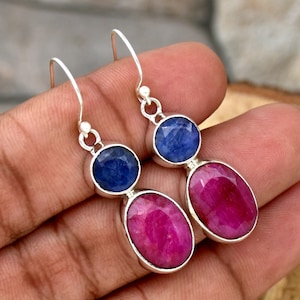Peut inclure: Une paire de boucles d'oreilles pendantes en argent avec une saphir bleu et une rubis rose. Les boucles d'oreilles ont un design en forme de goutte avec un saphir bleu rond en haut et un rubis rose ovale en bas.