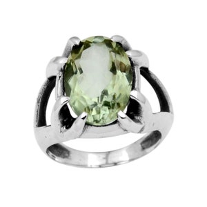 Puede incluir: Un anillo de plata con una gran piedra preciosa ovalada de color verde pálido. La piedra está engastada en un engaste calado detallado con detalles curvos. El anillo tiene un acabado pulido, que resalta la claridad y el color de la piedra.