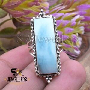 May include: A rectangular silver ring featuring a light blue gemstone with a marbled appearance. The ring has a detailed, textured border and decorative accents at the top and bottom. The ring is set against a blurred floral background.