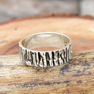 Op de afbeelding: Een zilveren ring met een getextureerd ontwerp dat lijkt op schors. De ring is breed en heeft een gladde binnenband.