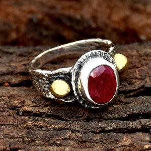 May include: A silver ring with a red gemstone and gold accents. The ring has a textured band and a bezel setting for the gemstone.
