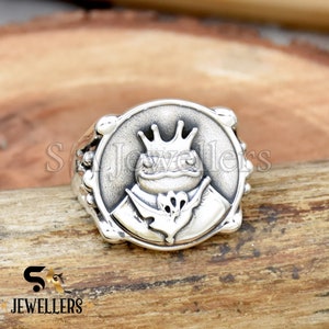 Könnte beinhalten: Ein silberner Ring mit einem detaillierten Design einer Kröte, die eine Krone trägt. Die Kröte hält eine Blume in der Hand. Der Ring ist in einer runden Fassung gefasst und hat ein strukturiertes Band.