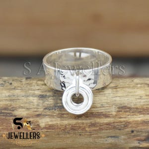 May include: A silver ring with a hammered texture and a dangling circular pendant. The ring is displayed on a wooden surface. The text "S A Jewellers" is visible on the ring and below it. The ring is a piece of jewelry.