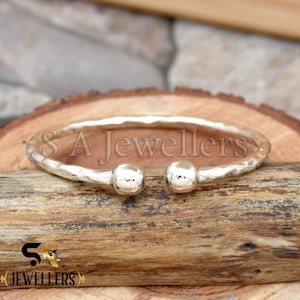 Pode incluir: Um bracelete de prata com acabamento martelado e duas pontas redondas. O bracelete está sobre uma superfície de madeira.
