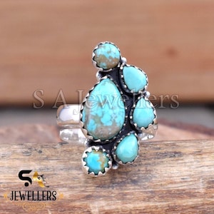 Può includere: Un anello d'argento con un gruppo di pietre turchesi di varie forme. L'anello ha un'incastonatura scura e ossidata che contrasta con le pietre blu-verdi brillanti. L'anello è appoggiato su una superficie di legno.