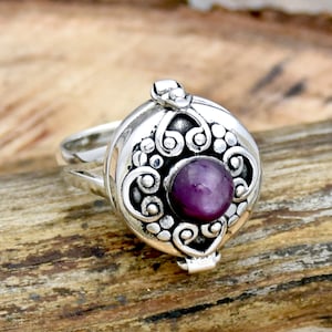 May include: A silver ring with an ornate design and a purple gemstone in the center. The ring is a locket that opens to reveal a small compartment.