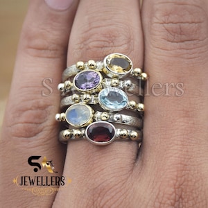 May include: Stackable sterling silver rings with oval gemstones in various colors: yellow, purple, blue, white, and red. Each ring has a silver band with gold-colored accents. The rings are displayed on a person's finger.