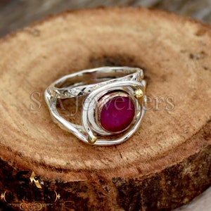May include: A silver ring with a pink gemstone set in a gold bezel. The ring has a unique design with two bands that wrap around the gemstone.