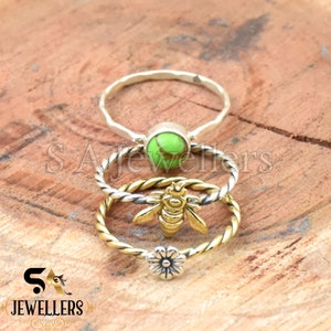 Pode incluir: Três anéis em uma superfície de madeira. O anel superior é prateado com uma pedra verde. O anel do meio é prateado e dourado com uma abelha. O anel inferior é dourado com uma flor. Os anéis são da SA Jewellers.