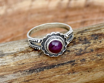 Puede incluir: Un anillo de plata con una piedra preciosa redonda de color rojo intenso engastada en un bisel en forma de flor. El anillo tiene una banda detallada con un diseño texturizado. El anillo se muestra sobre una superficie de madera.