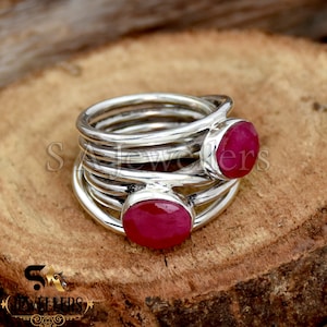 Puede incluir: Un anillo de plata de múltiples capas con dos piedras preciosas ovaladas de color rojo intenso. El diseño del anillo presenta múltiples bandas de plata, creando un efecto apilado. Las piedras preciosas están engastadas en la parte superior e inferior del anillo.