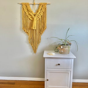 May include: A mustard yellow macrame wall hanging with a wooden dowel at the top. The hanging is in a diamond shape with a tassel at the bottom. The hanging is on a light grey wall next to a white bedside table with a drawer and a door.