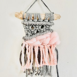 Puede incluir: Un tapiz de macramé gris y rosa con una rama de madera en la parte superior y una larga franja en la parte inferior.