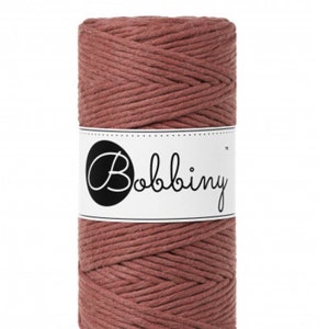 Puede incluir: Un carrete de cordón de macramé color rosa empolvado. El cordón está bien enrollado y envuelto con una etiqueta blanca con la palabra "Bobbiny" en una fuente cursiva. El cordón parece estar hecho de un material suave y texturizado.