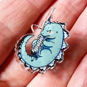 Könnte beinhalten: Ein blauer und weißer Cartoon-Dinosaurier mit stacheligem Rücken, der ein weiß-blau gestreiftes Objekt im Maul hält. Der Dinosaurier hat ein rotes Herz über seinem Kopf. Das Bild ist ein transparenter Acryl-Anhänger.