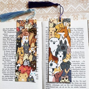 Könnte beinhalten: Zwei illustrierte Lesezeichen mit farbenfrohen Tiermotiven. Eines zeigt Katzen, das andere Hunde. Jedes Lesezeichen hat eine Quaste. Die Lesezeichen befinden sich in einem offenen Buch.