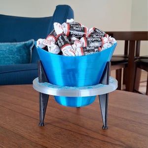 May include: A blue 3D-printed candy dish filled with Tootsie Rolls. The dish has a flared bowl shape and sits on a white and black stand with three legs. The candy dish is on a wooden table.
