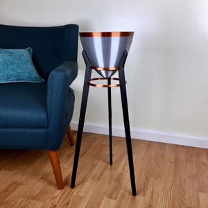 May include: A modern, silver metal planter with a copper rim and accents, supported by a black tripod base. The planter has a conical shape and is designed for indoor use. The overall height is approximately 36 inches.