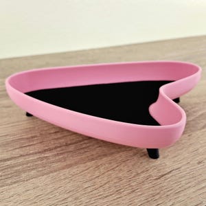 May include: A pink heart-shaped tray with a black interior and three black legs. The tray is made of a smooth material and sits on a wooden surface. The tray's design is modern and minimalist.