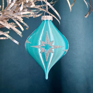 May include: A turquoise teardrop-shaped Christmas ornament with a silver glitter starburst design. The ornament has a silver cap and is hanging from a silver string. The background is a solid teal color.