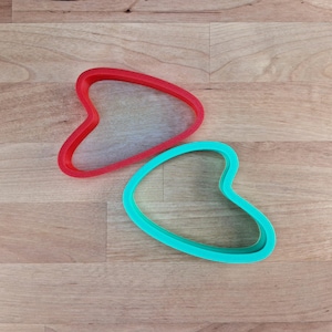 Peut inclure: Deux emporte-pièces colorés en forme de boomerang ou de cœur stylisé. L'un est rouge vif et l'autre est turquoise vif. Ils sont en plastique et reposent sur une surface en bois brun clair.